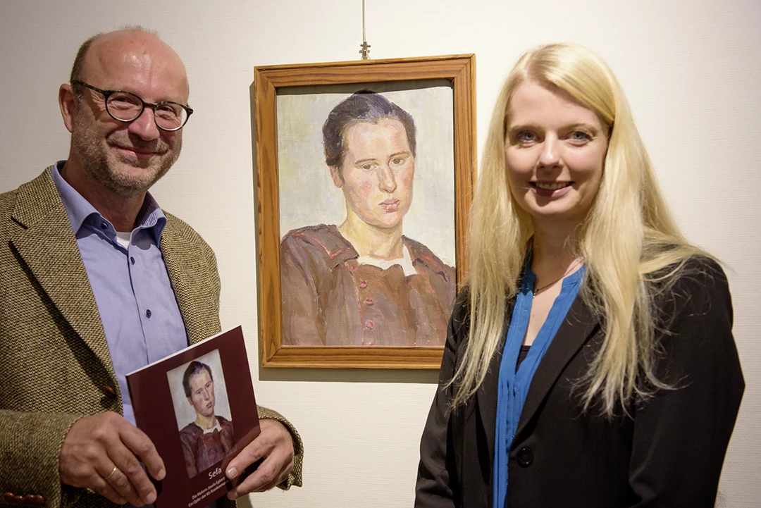 Ein Mann und eine Frau stehen vor einem Gemälde und lächeln in die Kamera.