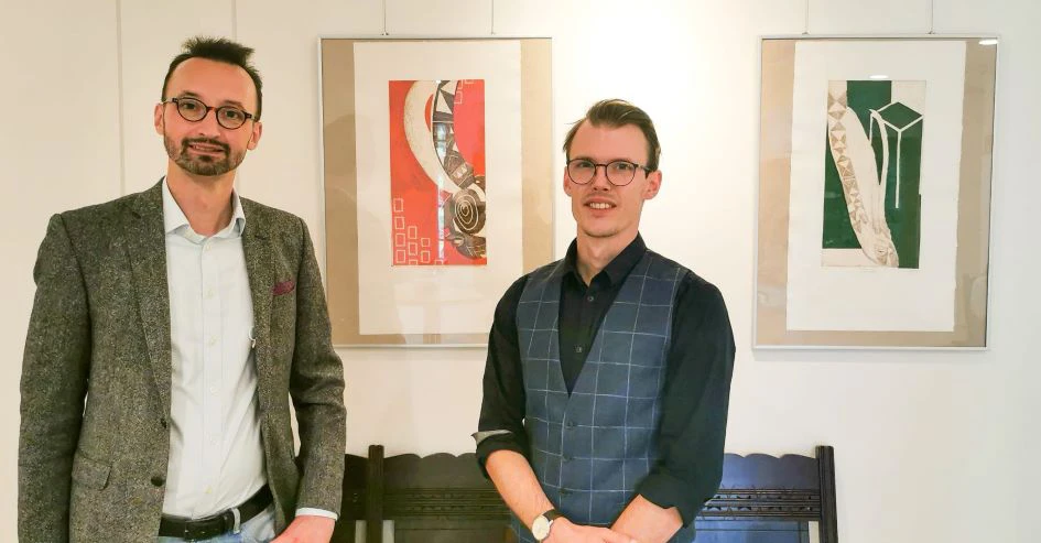 Zwei Männer stehen vor zwei Bilderrahmen und lächeln in die Kamera.