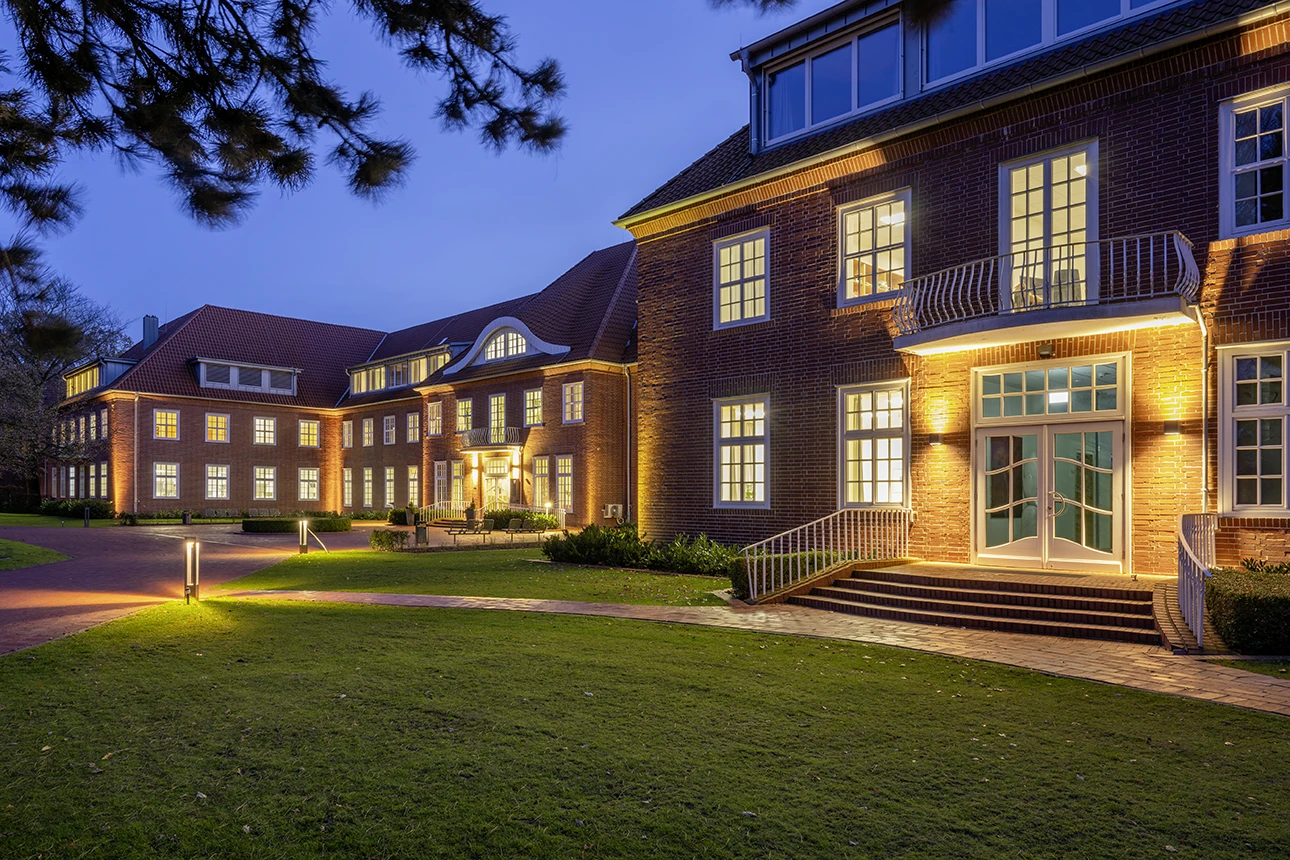 Seitlicher Blick auf das Gebäude der Katholischen Akademie Stapelfeld bei abendlicher Dämmerung. Im Gebäude und außen sind alle Lichter an.