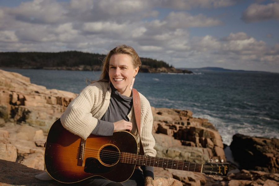 Carolline Cotter mit ihrer Gitarre auf Steinen sitzend vor einem Gewässer