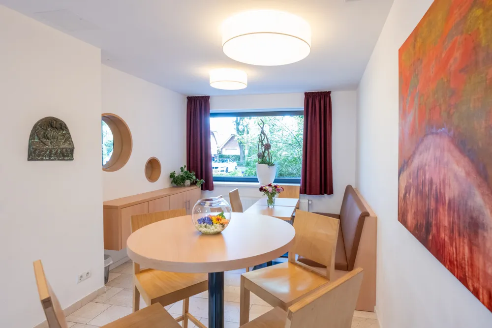 Im Aufenthaltsraum des Haus Emmaus steht im Vordergrund ein runder Tisch mit 4 Stühlen. Dahinter ist an der rechten Seite eine Sitzbank mit zwei kleinen Tischen davor und an der linken Wand hängt ein Sideboard.