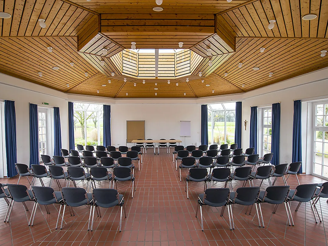 In der Aula der Katholischen Akademie Stapelfeld stehen mehrere Stuhlreihen mit Blick nach vorne, wo zwei Tische mit insgesamt vier Stühlen stehen. Dahinter stehen eine Pinnwand und ein Whiteboard.