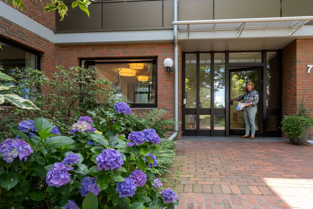 Blick auf den Eingang des Haus Emmaus, eine Frau steht in der Tür mit Blättern in der Hand. Im Vordergrund in der linken Ecke ist eine lilafarbene Pflanze.