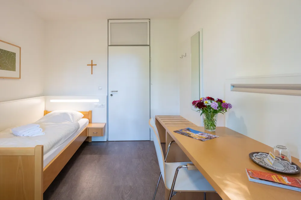 Ein Ein-Bett-Zimmer ist eingerichtet mit einem Schreibtisch, einem Einzelbett, einem kleinem Schrank und einem Nachtschrank. An der weißen Wand über dem Bett hängt ein Kreuz.