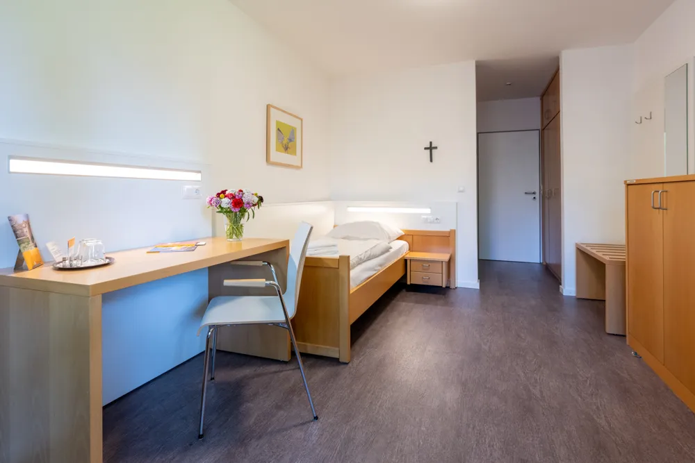 Ein Ein-Bett-Zimmer ist eingerichtet mit einem Schreibtisch, einem Einzelbett, einem kleinem Schrank und einem Nachtschrank. An der weißen Wand über dem Bett hängt ein Kreuz.