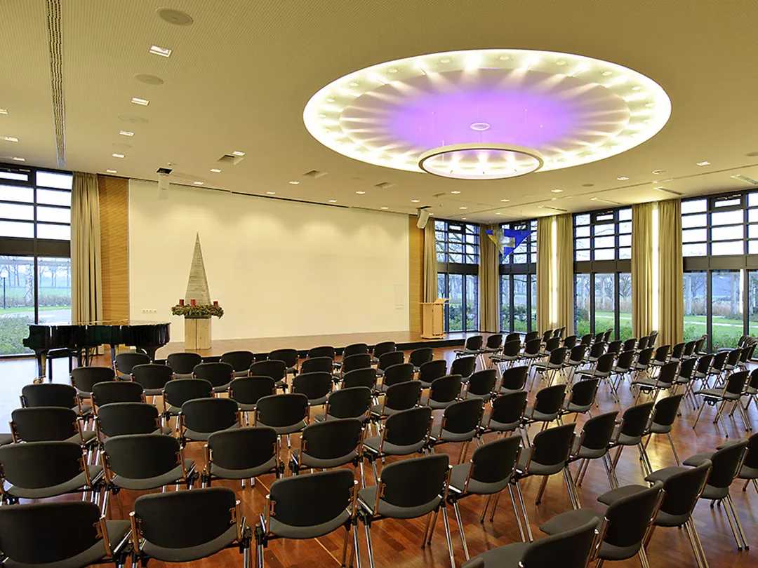 Im Forum der Katholischen Akademie Stapelfeld stehen viele leere Stuhlreihen hintereinander. Im Hintergrund ist eine leere Bühne zu sehen.