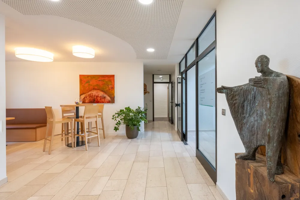 Im Foyer des Haus Emmaus stehen ein Stehtisch mit 4 Hochstühlen sowie eine Bank und neben dem Eingang eine mittelgroße Bronzestatue.