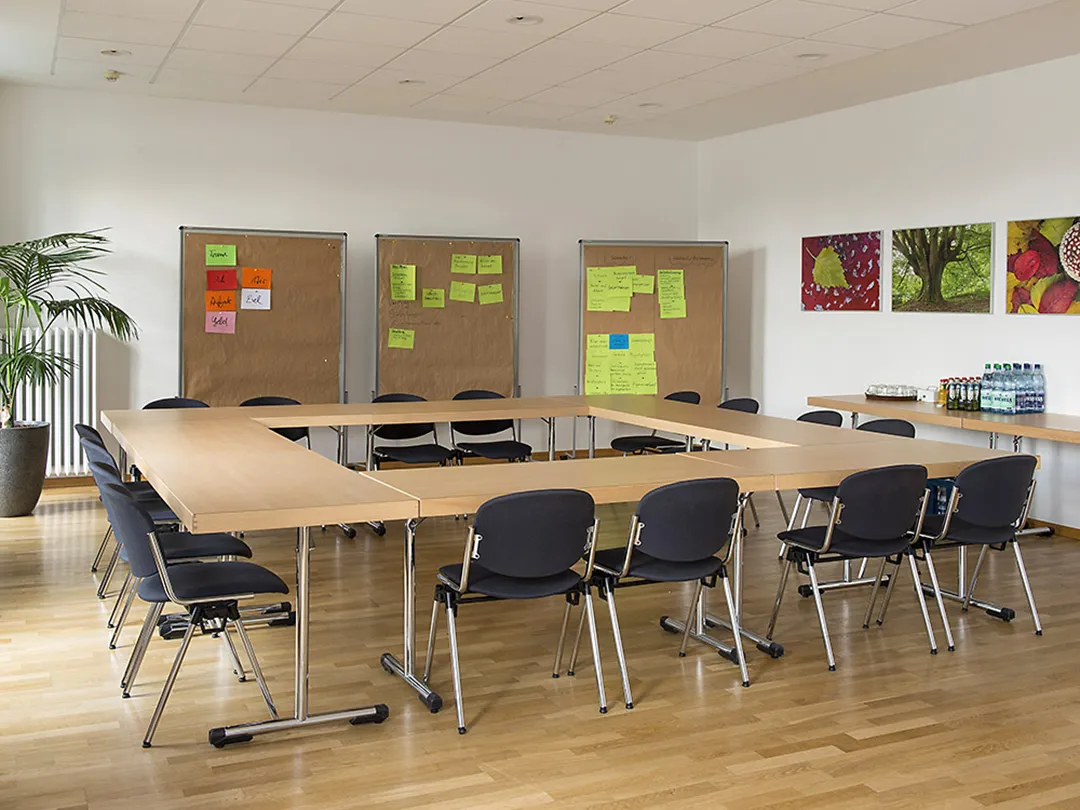 In einem Seminarraum stehen acht Tische in einem Kreis angeordnet. Im Hintergrund stehen drei Pinnwände mit Rollen, an denen beschriebene, bunte Zettel hängen.