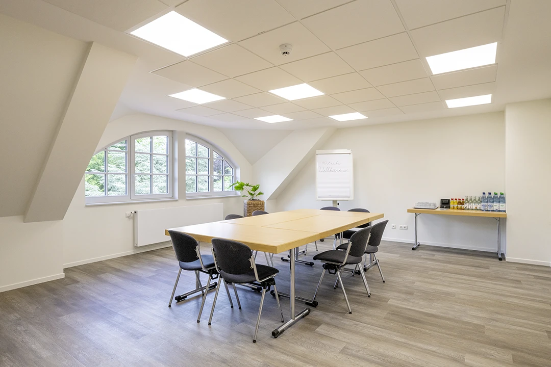 In einem hellen Seminarrum stehen vier Tische zu einem Rechteck angeordnet mitten im Raum mit Stühlen versehen. Im Hintergrund steht ein Tisch mit Getränken.