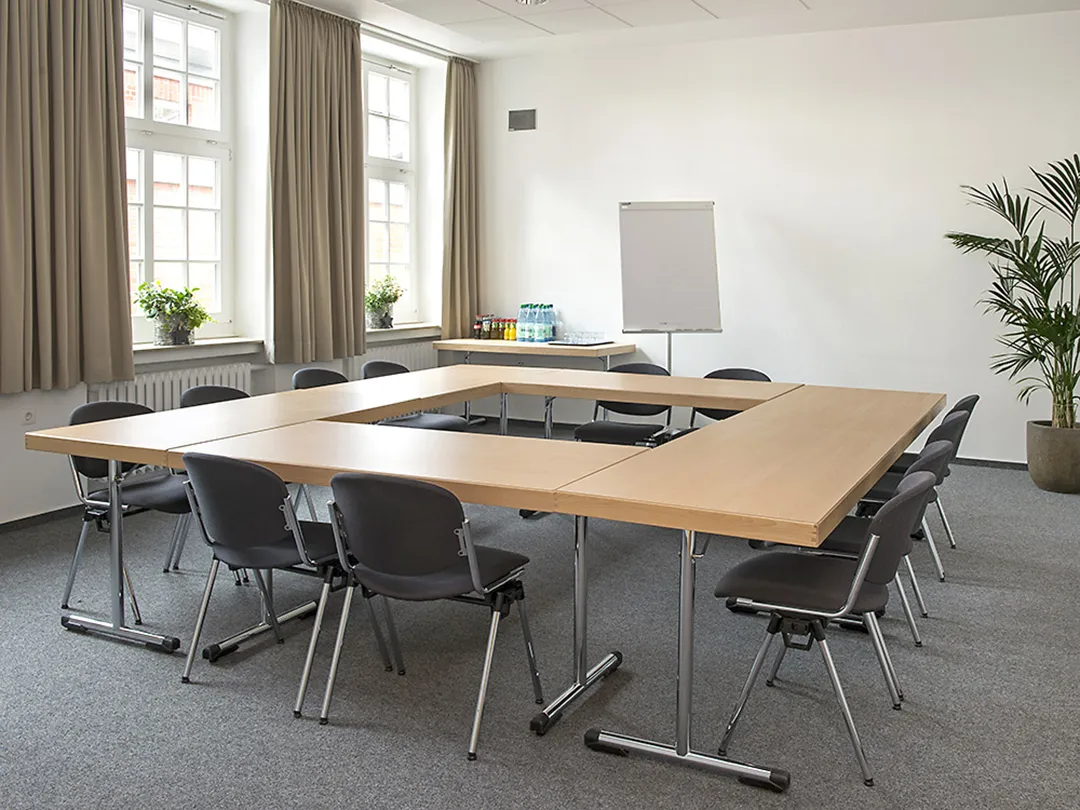 In einem Seminarraum mit Teppichboden und beigen Vorhängen stehen sechs Tische mit Stühlen zu einem O angeordnet.