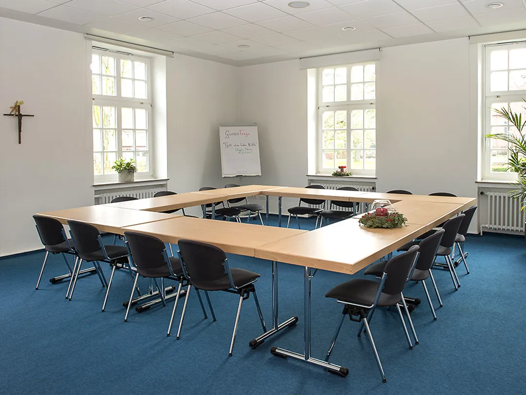 In einem Seminarraum mit Teppichboden stehen acht Tische in einem O angeordnet von Stühlen umgeben. Im Hintergrund steht ein beschriebenes Whiteboard.