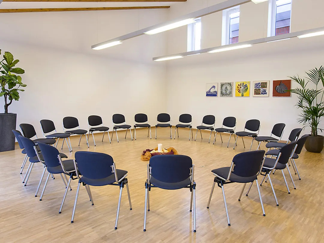 In einem Seminarraum mit Parkettboden stehen Stühle in einem Stuhlkreis. In der Mitte steht eine Kerze umgeben von Tüchern auf dem Boden.