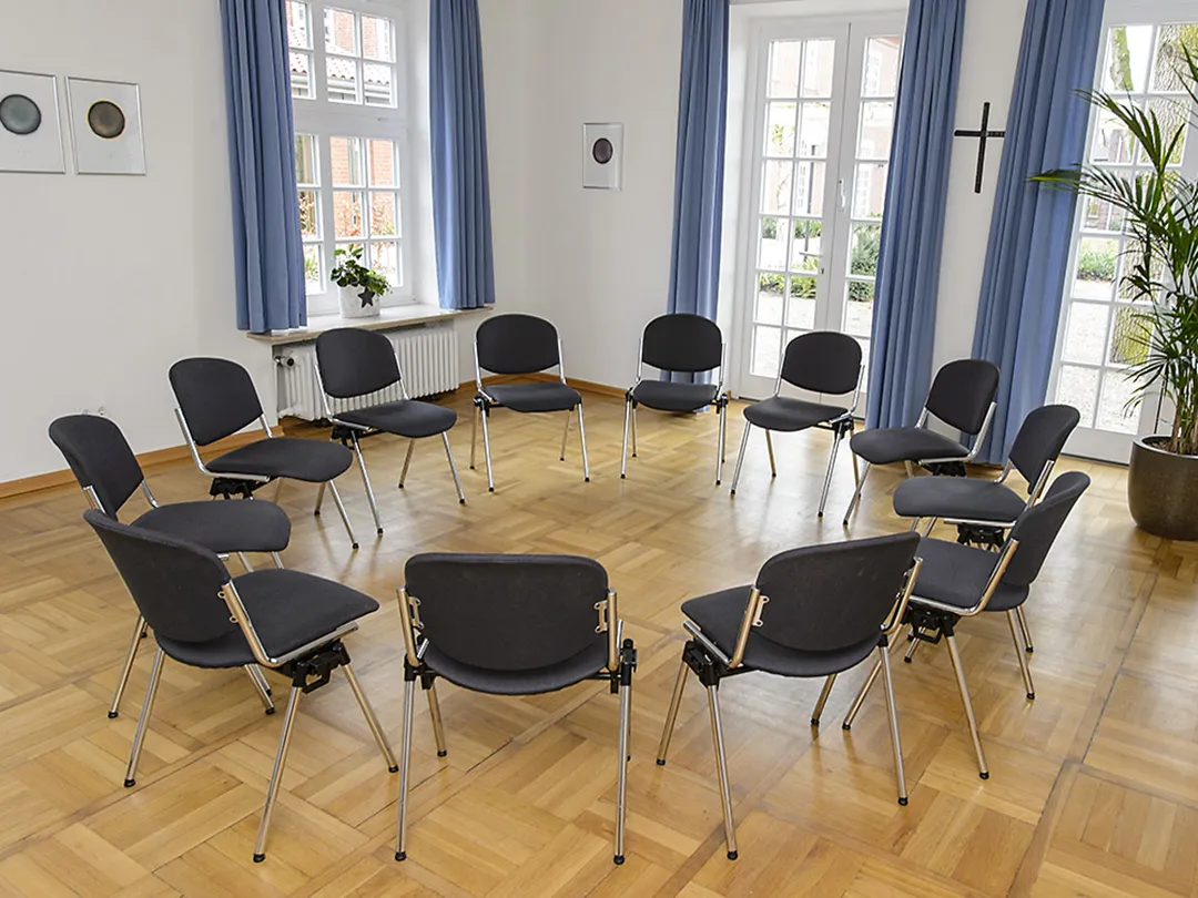 In einem Seminarraum mit Parkettboden und hellblauen Vorhängen steht ein Stuhlkreis.