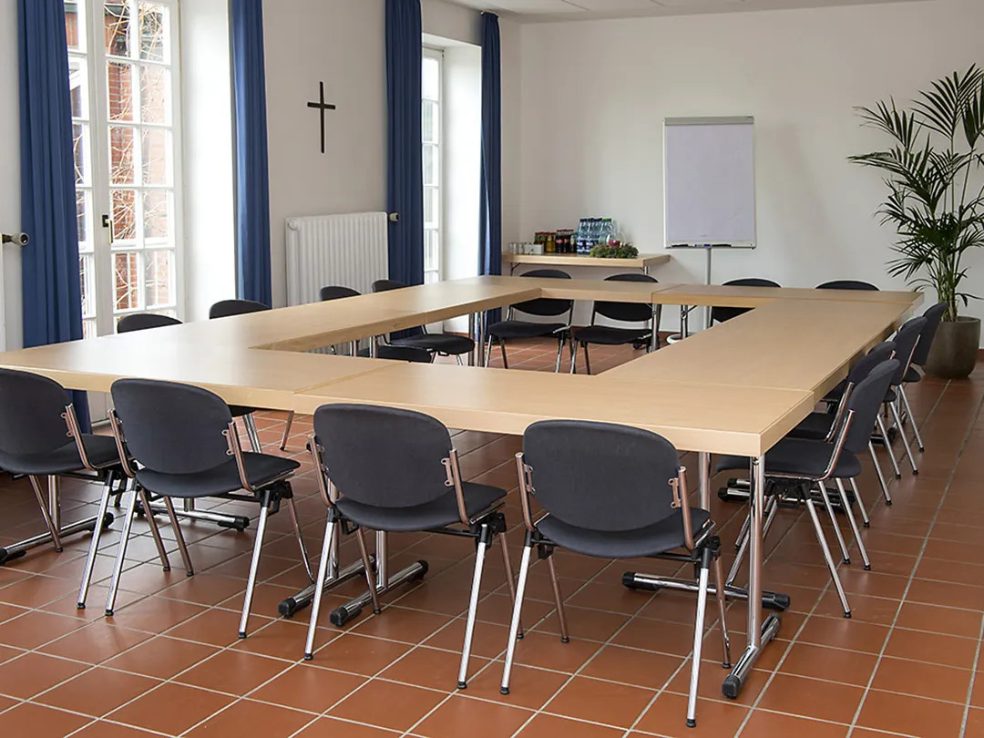 In einem Seminarraum mit roten Fliesen und blauen Gardinen stehen acht Tische zu einem O geformt mit Stühlen drumherum. Im Hintergrund steht ein leerer Block Papier an einer Pinnwand.