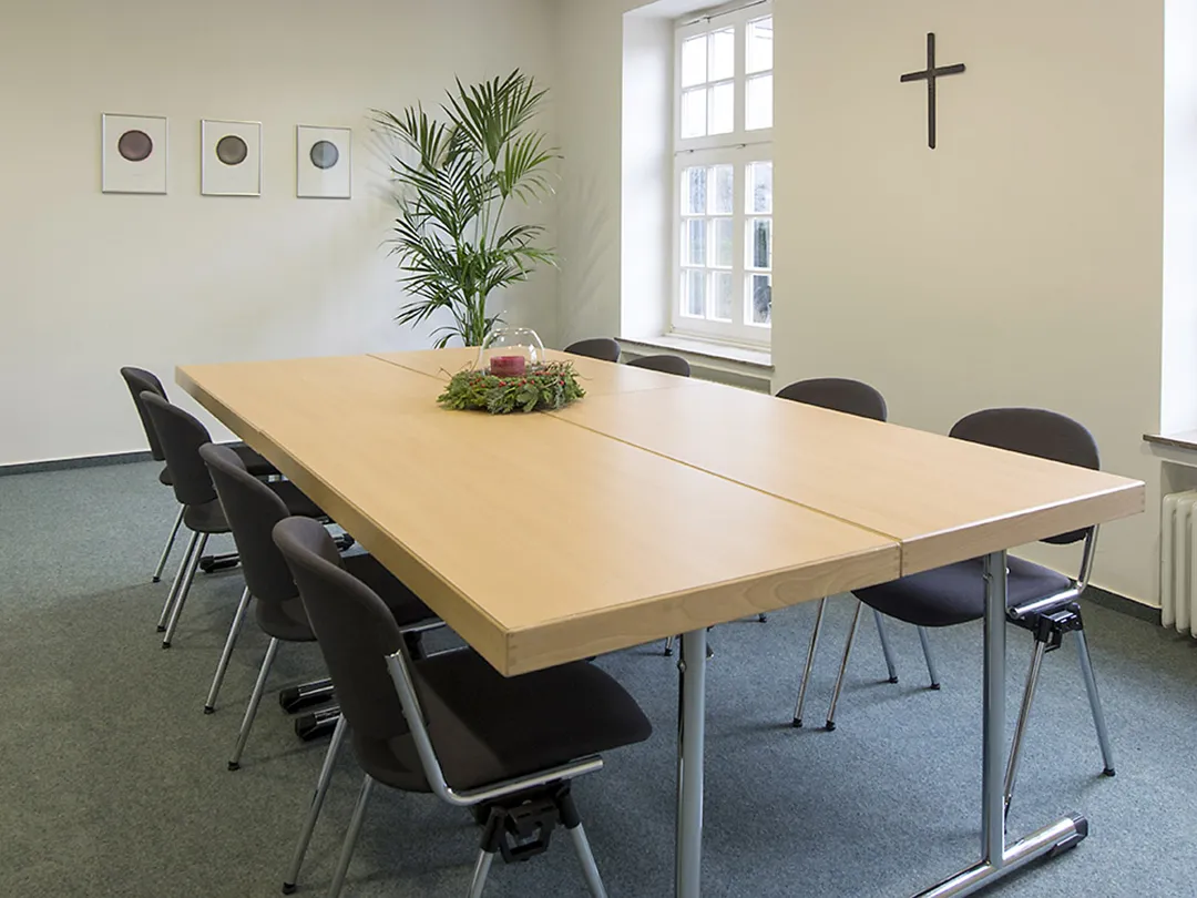 In einem Raum mit weißer Wand und Teppichboden stehen vier Tische zusammen als Rechteck mit Tischen drumherum. An der einen Wand hängt ein kleines Kreuz.