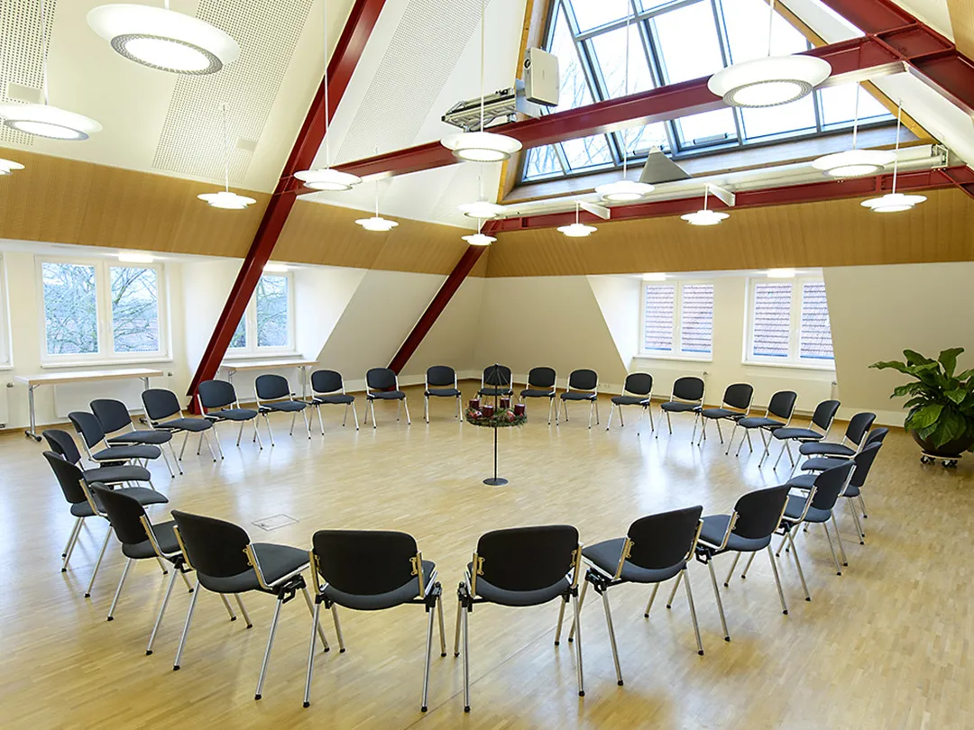 In einem Raum unter einem Dach stehen Stühle in einem Stuhlkreis, in der Mitte davon steht ein Adventskranz mit vier Kerzen. Im Hintergrund befinden sich Fenster.