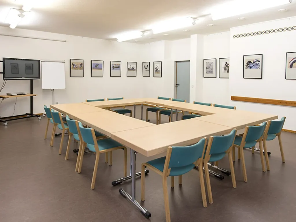 In einem Meeting-Raum stehen Tische in einem O mit Stühlen versehen in der Mitte. An den Wänden hängen Bilder von Vögeln in einer Reihe