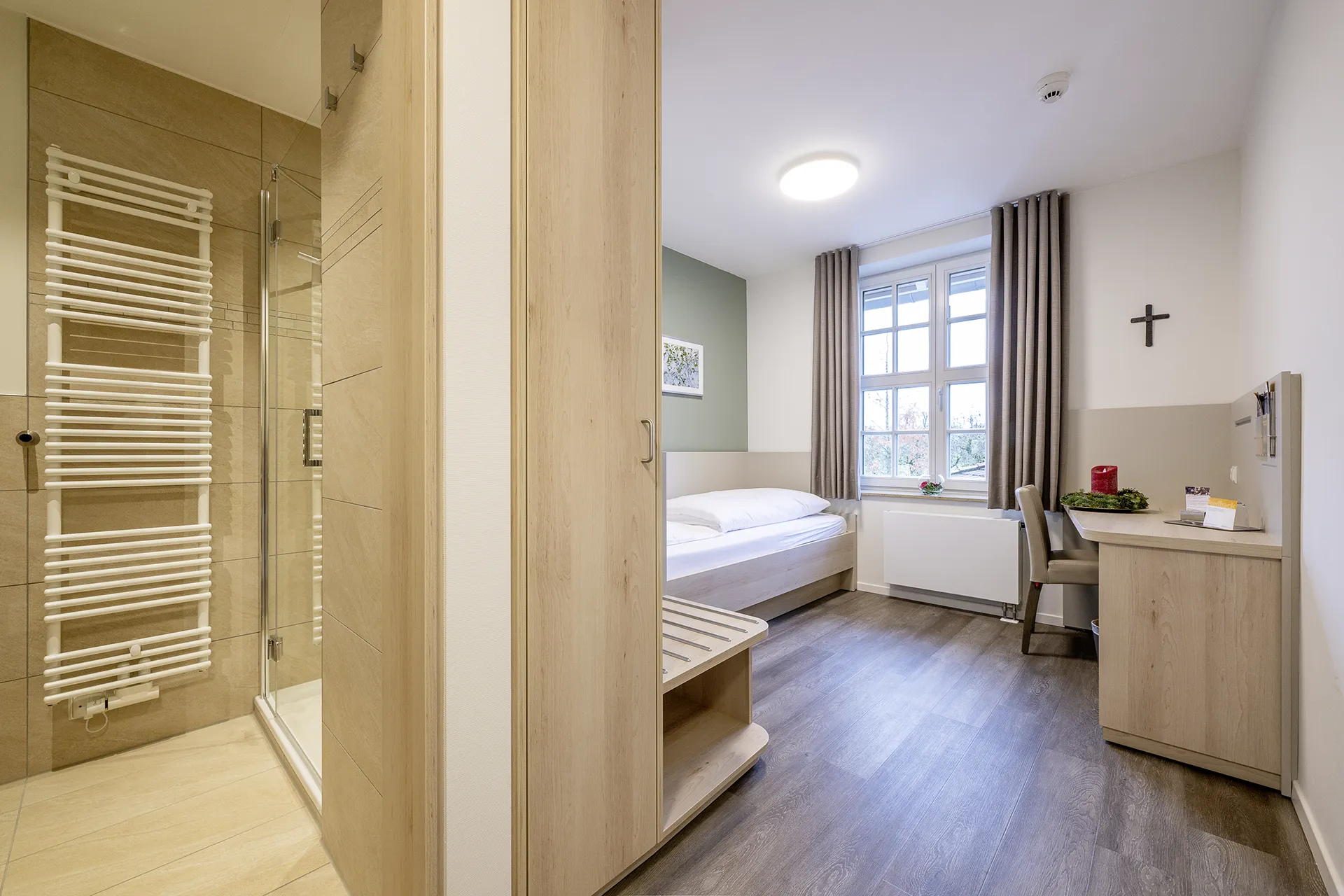 Blick in ein Zimmer, auf der linken Seite ist eine Duschecke mit einer Wandheizung. Der Rest des Raumes ist mit einem Bett, einem Fenster und einem Schreibtisch an den Wänden ausgestattet.