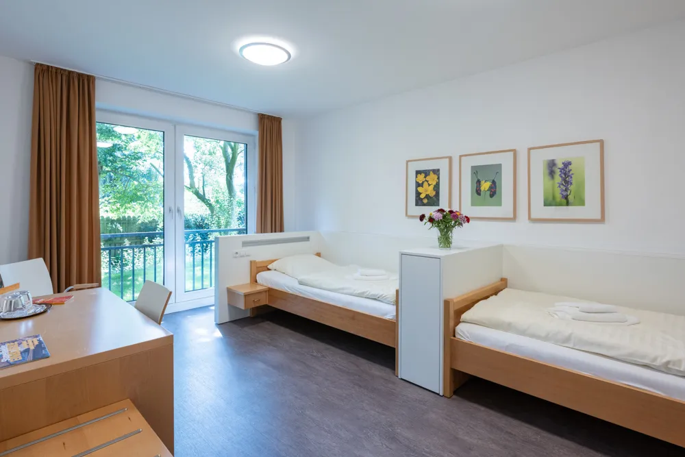 Zwei Betten stehen Fuß an Fuß getrennt durch ein Einschubregal in einem Raum an der rechten Wandseite mit einem Doppelfenster, das bis zum Boden reicht. An der linken Seite steht ein Schreibtisch mit einem angeschobenen Stuhl.